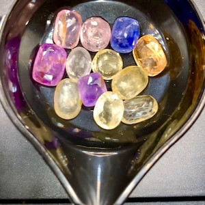 May include: A collection of colorful gemstones in various shapes and sizes, including pink, purple, yellow, and blue, are displayed in a black, oval-shaped container. The gemstones are on a scale, suggesting they are being weighed or measured.