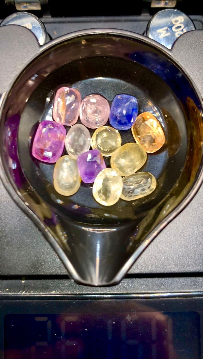 May include: A collection of colorful gemstones in various shapes and sizes, including pink, purple, yellow, and blue, are displayed in a black, oval-shaped container. The gemstones are on a scale, suggesting they are being weighed or measured.