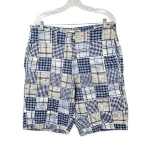 May include: Patchwork shorts in shades of blue, white, and gray, hanging on a white hanger. The shorts feature a button closure and a mix of plaid patterns. The shorts are made of a lightweight fabric.