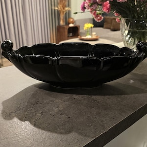 May include: A glossy black decorative bowl with a scalloped rim and two ornate handles. The bowl sits on a dark countertop. In the background, a vase of pink flowers and a window with curtains are visible.