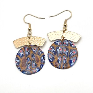 Peut inclure: Une paire de boucles d'oreilles pendantes dorées avec un pendentif rond à motifs bleus et bruns et une barre dorée martelée au-dessus du cercle.