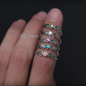 K&ouml;nnte beinhalten: F&uuml;nf silberne Ringe mit Engelsfl&uuml;gel-Designs. Jeder Ring hat einen andersfarbigen Edelstein: gr&uuml;n, wei&szlig;, rosa, blau und Opal.
