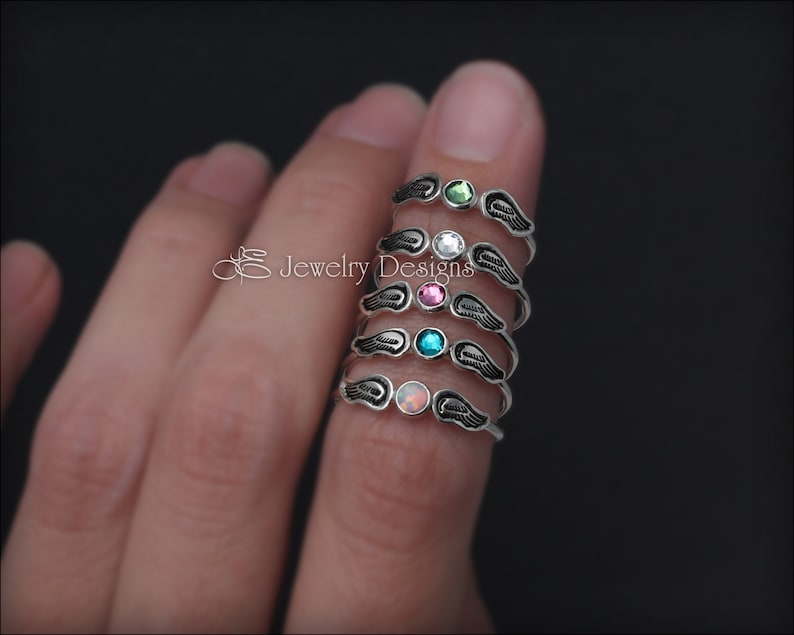 K&ouml;nnte beinhalten: F&uuml;nf silberne Ringe mit Engelsfl&uuml;gel-Designs. Jeder Ring hat einen andersfarbigen Edelstein: gr&uuml;n, wei&szlig;, rosa, blau und Opal.