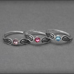 K&ouml;nnte beinhalten: Zwei silberne Ringe mit Engelsfl&uuml;gel-Designs und bunten Edelsteinen. Der obere Ring hat einen hellblauen Edelstein und der untere Ring einen rosafarbenen Edelstein.