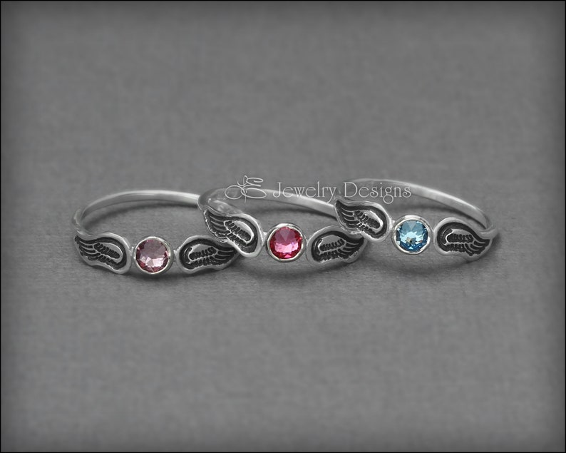K&ouml;nnte beinhalten: Zwei silberne Ringe mit Engelsfl&uuml;gel-Designs und bunten Edelsteinen. Der obere Ring hat einen hellblauen Edelstein und der untere Ring einen rosafarbenen Edelstein.