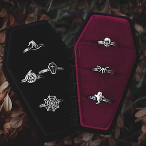 May include: A collection of silver-toned Halloween-themed rings displayed in open coffin-shaped ring boxes. The rings feature designs such as a witch's hat, a pumpkin, a coffin, a spider web, a skull, a spider, and a ghost.