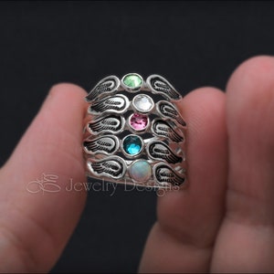 K&ouml;nnte beinhalten: F&uuml;nf silberne Ringe mit Engelsfl&uuml;gel-Designs. Jeder Ring hat einen andersfarbigen Edelstein: gr&uuml;n, klar, rosa, blau und wei&szlig;.