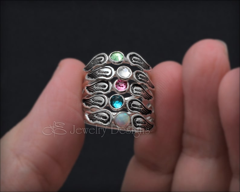 K&ouml;nnte beinhalten: F&uuml;nf silberne Ringe mit Engelsfl&uuml;gel-Designs. Jeder Ring hat einen andersfarbigen Edelstein: gr&uuml;n, klar, rosa, blau und wei&szlig;.
