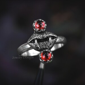 May include: A silver ring featuring a stylized design of open lips with sharp teeth, flanked by two red gemstone accents. The ring has a dark, gothic aesthetic, with the words "Jewelry Designs" visible.
