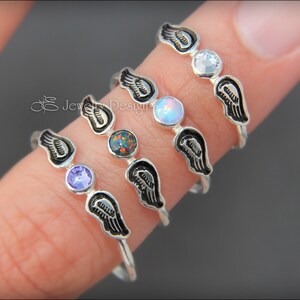 K&ouml;nnte beinhalten: Vier Silberringe mit Engelsfl&uuml;gel-Designs und verschiedenen farbigen Edelsteinen. Die Ringe sind auf einem Finger &uuml;bereinander gelegt. Die Edelsteine sind lila, Opal, schwarz und klar.