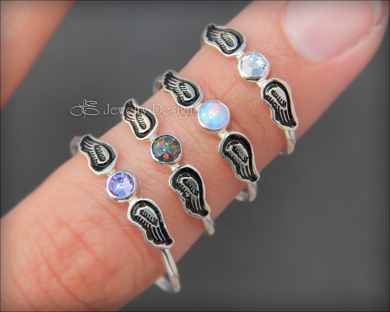 K&ouml;nnte beinhalten: Vier Silberringe mit Engelsfl&uuml;gel-Designs und verschiedenen farbigen Edelsteinen. Die Ringe sind auf einem Finger &uuml;bereinander gelegt. Die Edelsteine sind lila, Opal, schwarz und klar.