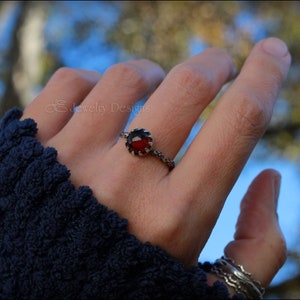 May include: A silver ring with a red gemstone set in a crown-like design. The ring is worn on the index finger of a hand.