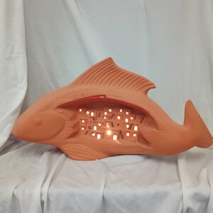 May include: A terracotta-colored fish-shaped lamp with a hollowed-out center. The interior reveals a series of small, illuminated squares, creating a warm, inviting glow. The lamp's design features detailed fins and a tail, adding to its artistic appeal.