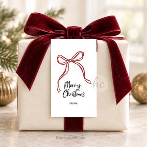 May include: A white gift tag featuring a red line-art bow illustration, the black text Merry Christmas, and the word FROM followed by a colon. The tag is attached to a cream-colored gift box wrapped with a large, dark red velvet ribbon bow.