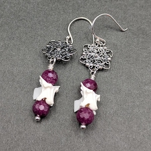 May include: A pair of drop earrings featuring silver-toned filigree connectors, each suspending a faceted deep purple bead above a white, bone-shaped spacer and a second matching purple bead, all attached to simple silver-toned ear wires.