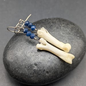 May include: A pair of dangle earrings featuring silver-toned metal hooks, each adorned with three faceted deep blue beads and a single off-white, smooth, elongated bone-shaped charm suspended at the bottom of the wire construction.