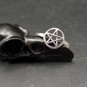 May include: A silver ring featuring a pentagram design within a circular frame. The ring is set against a dark background, highlighting its metallic sheen. The ring's design is a prominent feature, showcasing the intricate star pattern.