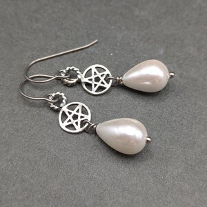 May include: A pair of drop earrings featuring silver-toned metal hooks, a circular charm with a cut-out five-pointed star pentagram design, and a single white teardrop-shaped faux pearl bead suspended from the bottom of each star charm.