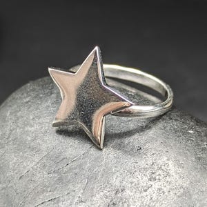 May include: A silver-colored ring featuring a five-pointed star design. The star is the focal point, with a polished, reflective surface. The ring band is thin and also silver, creating a simple yet elegant piece of jewelry.