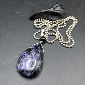May include: A teardrop-shaped pendant necklace with a black and purple stone, wire wrapped and suspended from a silver ball chain. A black, carved object rests above the necklace. The pendant is approximately 2 inches long.