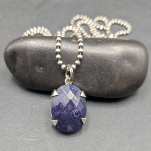 May include: A necklace featuring a faceted, oval-shaped deep blue gemstone pendant set in a four-prong silver-toned metal setting. The pendant hangs from a silver-toned ball chain, which is draped behind the stone in a circular pattern.
