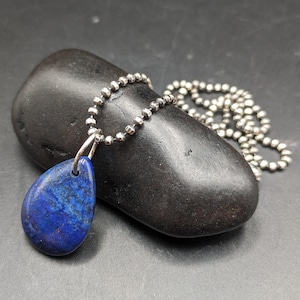May include: A teardrop-shaped lapis lazuli pendant necklace with a silver-colored chain. The deep blue stone has white and gold flecks. The necklace is displayed on a dark gray stone against a dark background.