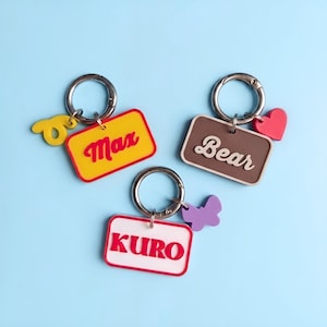 Puede incluir: Tres llaveros rectangulares con anillas de metal plateado. Una etiqueta amarilla dice Max con un dije de espiral amarillo. Una etiqueta marrón dice Bear con un corazón rojo. Una etiqueta blanca con borde rojo dice KURO con una mariposa morada.