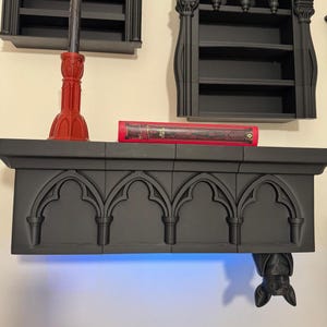 Könnte beinhalten: Ein schwarzes Regal im gotischen Stil mit gewölbten Details, einem roten Buch und einem roten Dekorationsgegenstand. Eine Fledermaus hängt von der Unterseite des Regals. Zwei schwarze Regale sind oben an der Wand befestigt.