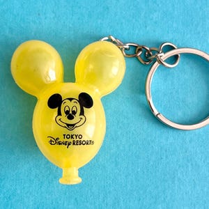 May include: Yellow Mickey Mouse-shaped keychain from Tokyo Disney Resort. The keychain features a smiling Mickey Mouse face and the Tokyo Disney Resort logo. The keychain is attached to a silver metal keyring.