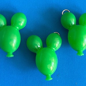 May include: Three bright green, Mickey Mouse-shaped charms with small silver hoops at the top. The charms have a glossy finish and are set against a solid blue background. These charms are likely used for jewelry or craft projects.