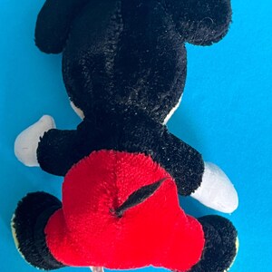 May include: A plush toy of a famous cartoon mouse, viewed from the back. The toy has black fur, large black ears, a red bottom, white gloves, and yellow feet. The toy is on a blue background.