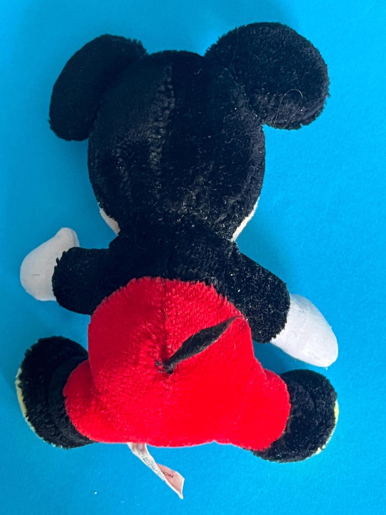 May include: A plush toy of a famous cartoon mouse, viewed from the back. The toy has black fur, large black ears, a red bottom, white gloves, and yellow feet. The toy is on a blue background.