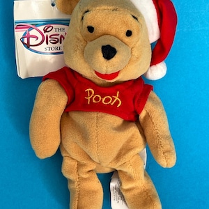 May include: A tan plush Winnie the Pooh bear wearing a red shirt with the name Pooh embroidered in yellow and a red and white Santa hat. The bear has a black nose and a red mouth. A white Disney Store tag is attached to the side of the bear.