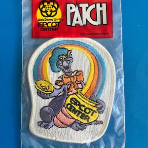 May include: An embroidered fabric patch featuring the purple dragon Figment from Epcot Center, holding a gold coin and a yellow pot with the text EPCOT CENTER, set against a rainbow. The patch is sealed in a clear plastic bag with a red header card.