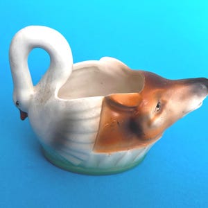 May include: A ceramic creamer shaped like a swan with a white body and a brown dog head. The swan neck forms the handle, while the dog head serves as the spout. The base features a subtle green tint, and the interior is a plain white finish.