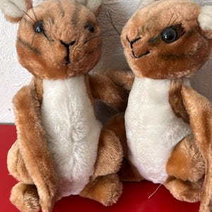 May include: Two plush toy squirrels with brown fur and white bellies. The squirrels have black eyes, whiskers, and small ears. They are sitting close together, with one slightly in front of the other. The background is a white wall and a red surface.