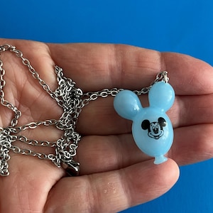 May include: A light blue Mickey Mouse balloon pendant necklace with a silver chain. The pendant features a black and white Mickey Mouse face. The necklace is displayed against a blue background.