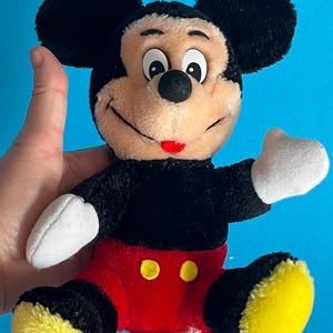 May include: A plush Mickey Mouse toy with black fur, large black ears, and a tan face. He wears red shorts with yellow buttons, white gloves, and yellow shoes. The toy is held against a blue background.