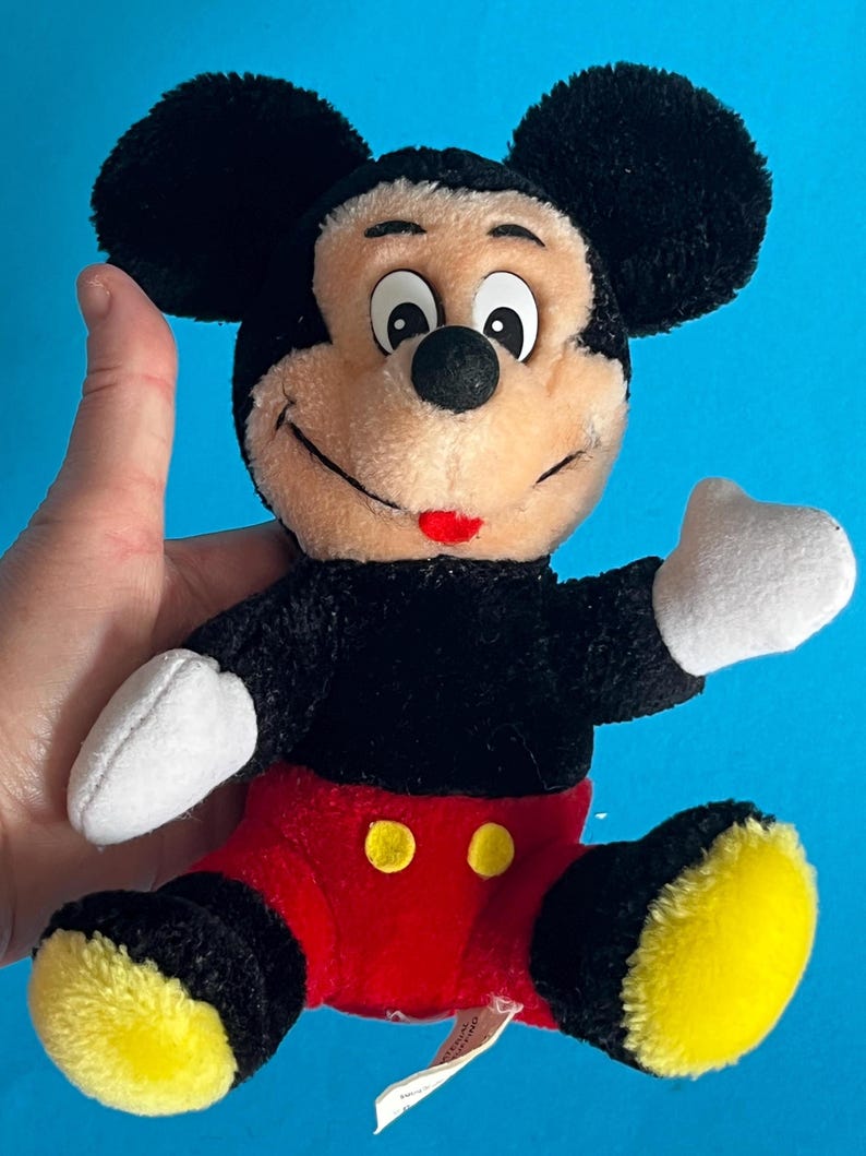 May include: A plush Mickey Mouse toy with black fur, large black ears, and a tan face. He wears red shorts with yellow buttons, white gloves, and yellow shoes. The toy is held against a blue background.