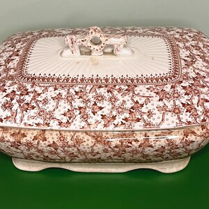 May include: A rectangular, antique serving dish with a lid. The dish features a white base with a brown floral pattern. The lid has a white center with a decorative handle and a brown border. The handles on the sides are also brown.