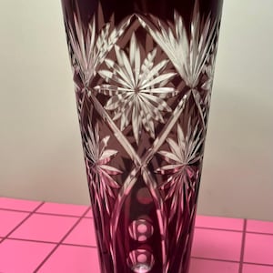 May include: A tall, burgundy-colored crystal vase with a detailed starburst and diamond pattern. The vase is approximately 10 inches tall and features a flared top and a series of circular designs near the base. The cut glass design reflects light.