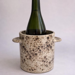 May include: A ceramic wine chiller with a speckled beige and brown pattern. A green wine bottle is inside the chiller. The chiller has two small handles on the sides. The background is white.