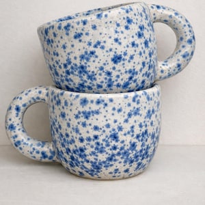 May include: Two ceramic mugs stacked on top of each other. The mugs are white with a speckled blue pattern. Each mug has a rounded handle. The mugs are designed for beverages.