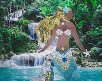 May include: A handcrafted mermaid figure with a blue tail, adorned with white lace, gold beads, and a large blue gem. The mermaid has blonde hair, daisy-shaped accents, and a starfish. The background features a waterfall and lush green foliage.