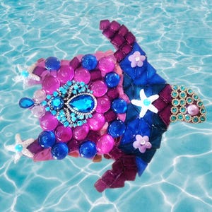 May include: A decorative mosaic turtle, crafted with a colourful array of glass pieces. The design incorporates blue, pink, and purple hues, embellished with jewels and starfish. The turtle is set against a blue water background.