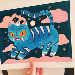 May include: An illustration of a blue cat with orange eyes, white fangs, and a bird wearing a hat. The cat has white stripes and is set against a dark teal background with pink clouds and gold stars. The artwork is signed in the bottom right corner.