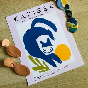 May include: A pink poster with the text "CATISSE THE MEWS DE MUSE." and a blue cat illustration with a yellow circle. The text "SAM. SEATTLE ART MUSEUM 1977" is at the bottom of the poster. There are three pairs of clay earrings in the image.