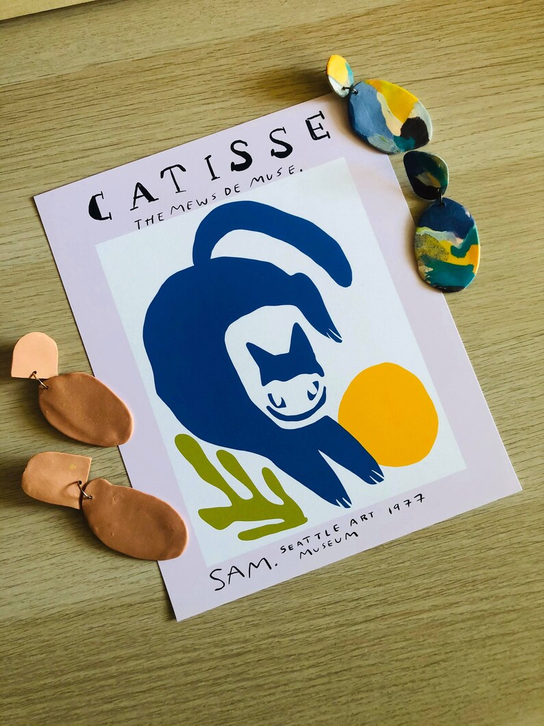 May include: A pink poster with the text "CATISSE THE MEWS DE MUSE." and a blue cat illustration with a yellow circle. The text "SAM. SEATTLE ART MUSEUM 1977" is at the bottom of the poster. There are three pairs of clay earrings in the image.