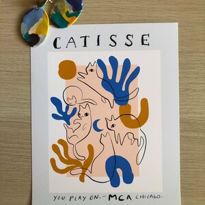 May include: A white poster with the text "CATISSE" in black lettering at the top. The poster features a colorful abstract design with three cats in a pink, blue, yellow, and orange color scheme. The text "YOU PLAY ON. - MCA CHICAGO." is at the bottom of the poster.