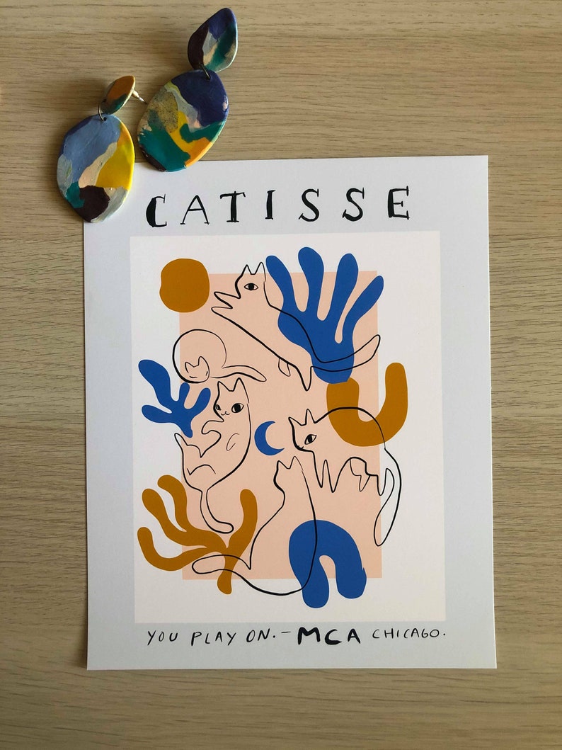 May include: A white poster with the text "CATISSE" in black lettering at the top. The poster features a colorful abstract design with three cats in a pink, blue, yellow, and orange color scheme. The text "YOU PLAY ON. - MCA CHICAGO." is at the bottom of the poster.
